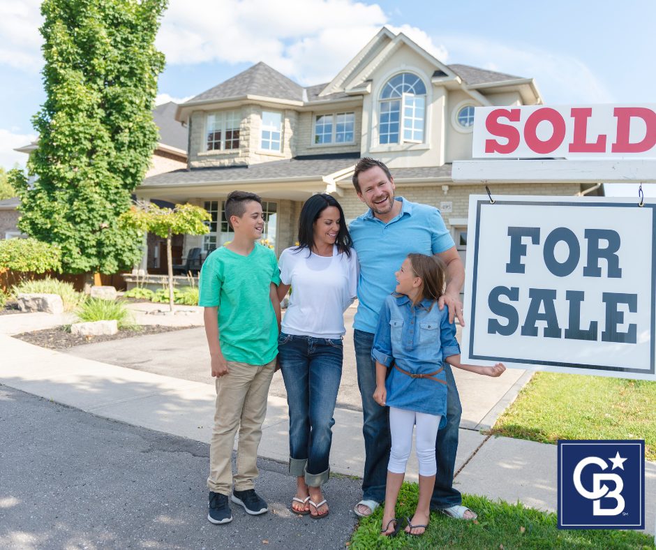 Smiling family in their front yard beside a “For Sale” sign with a “SOLD” rider—buying a home in South Metro Denver right now.